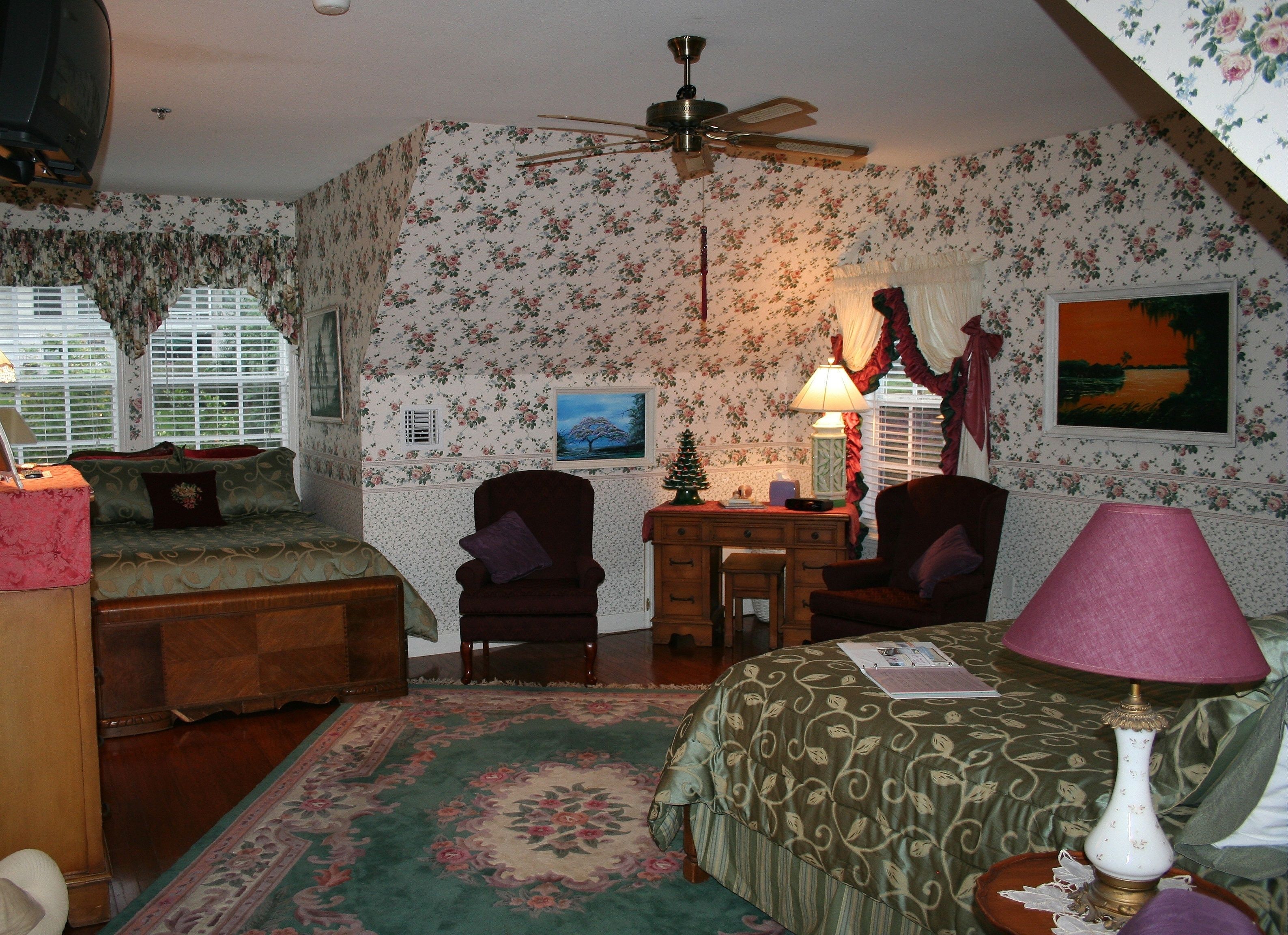Une chambre mansardée chaleureuse, avec un papier peint à motifs floraux, un ventilateur de plafond, un lit recouvert d’une courtepointe à motifs verts, deux fauteuils, un petit bureau en bois et une table de chevet avec une lampe.