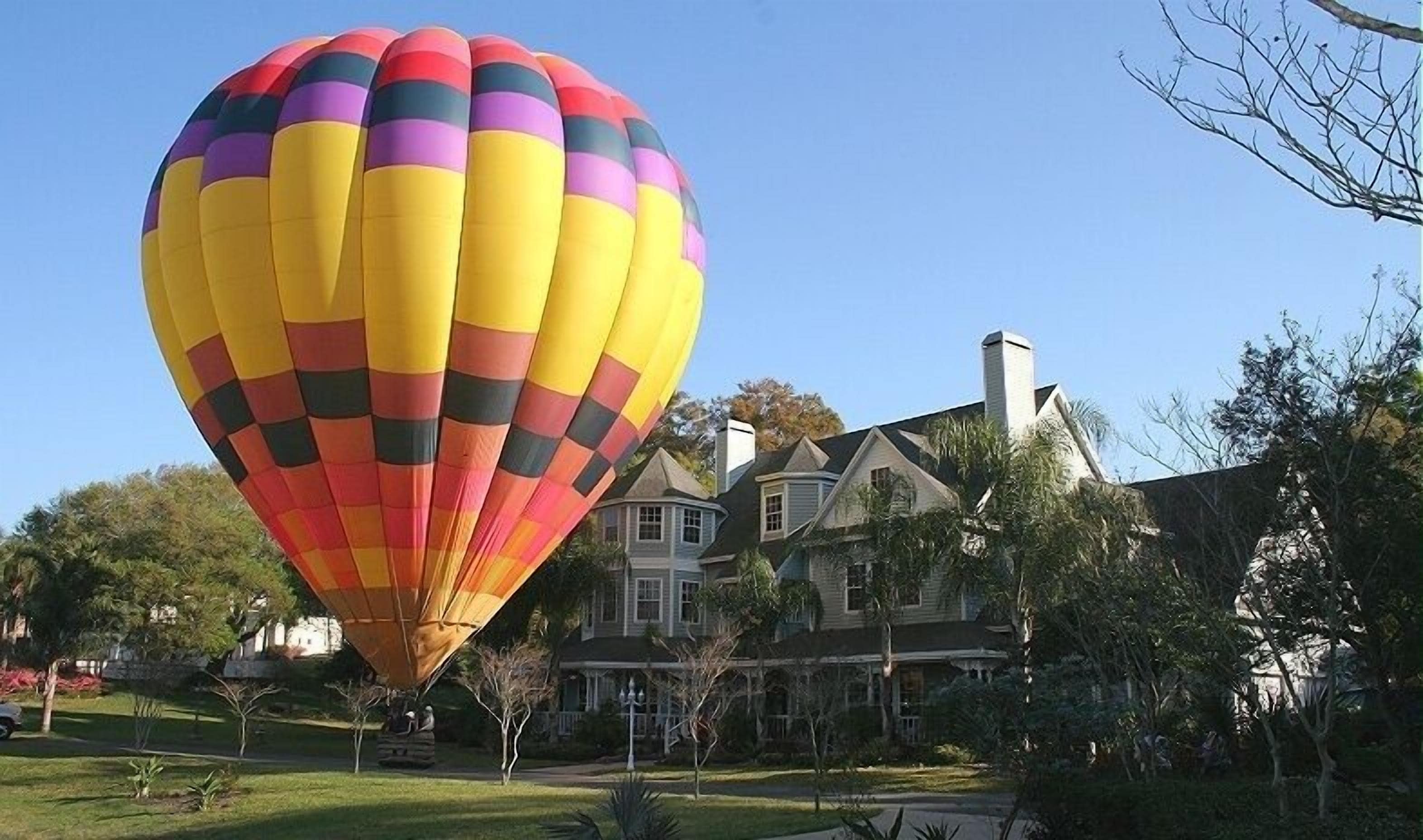 A hot air balloon with a multicolored pattern, parked in front of a large house with a chimney and surrounded by trees.