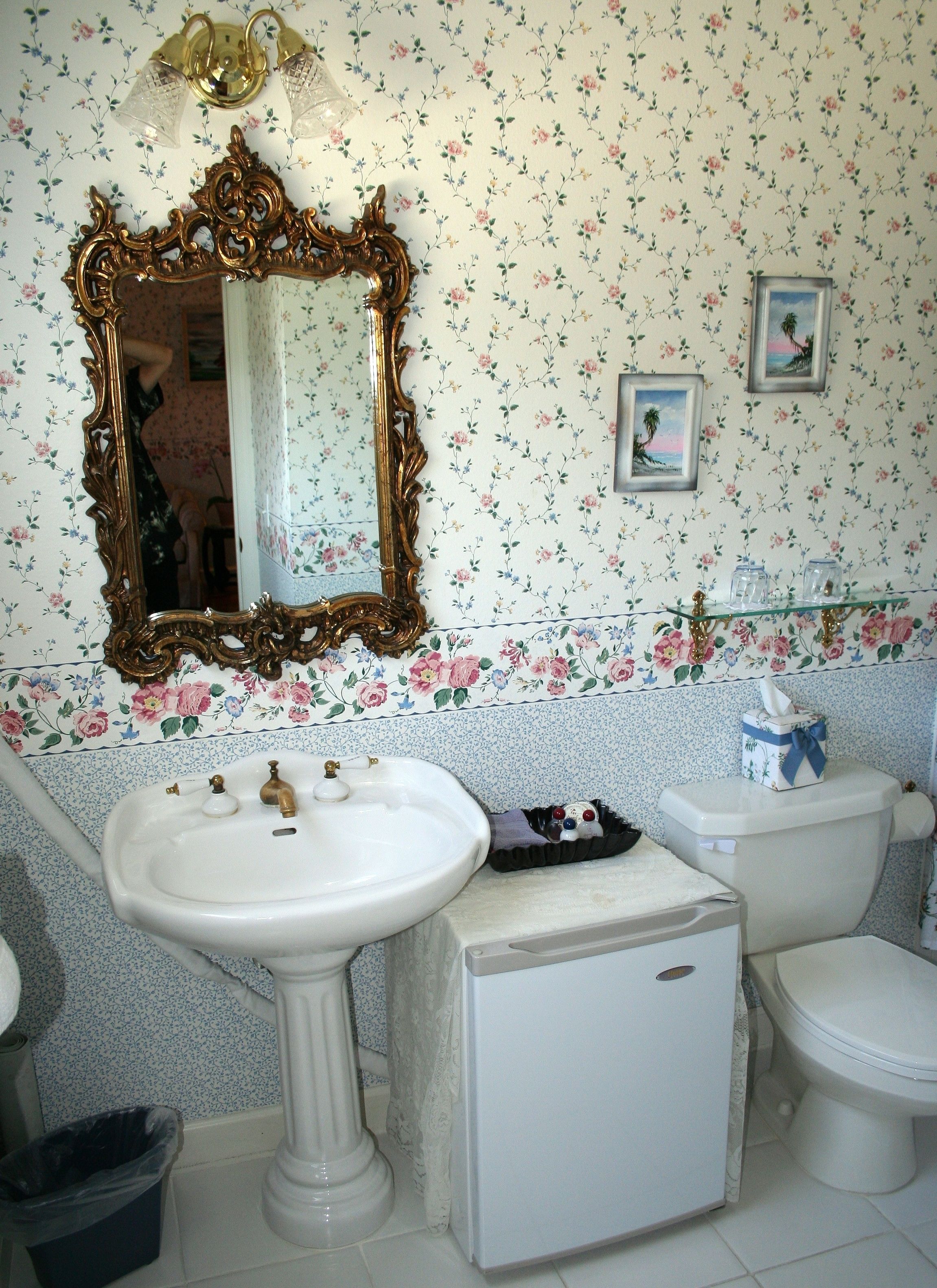 Un baño con papel tapiz floral, un lavamanos pedestal, un inodoro y un espejo con marco ornamentado.