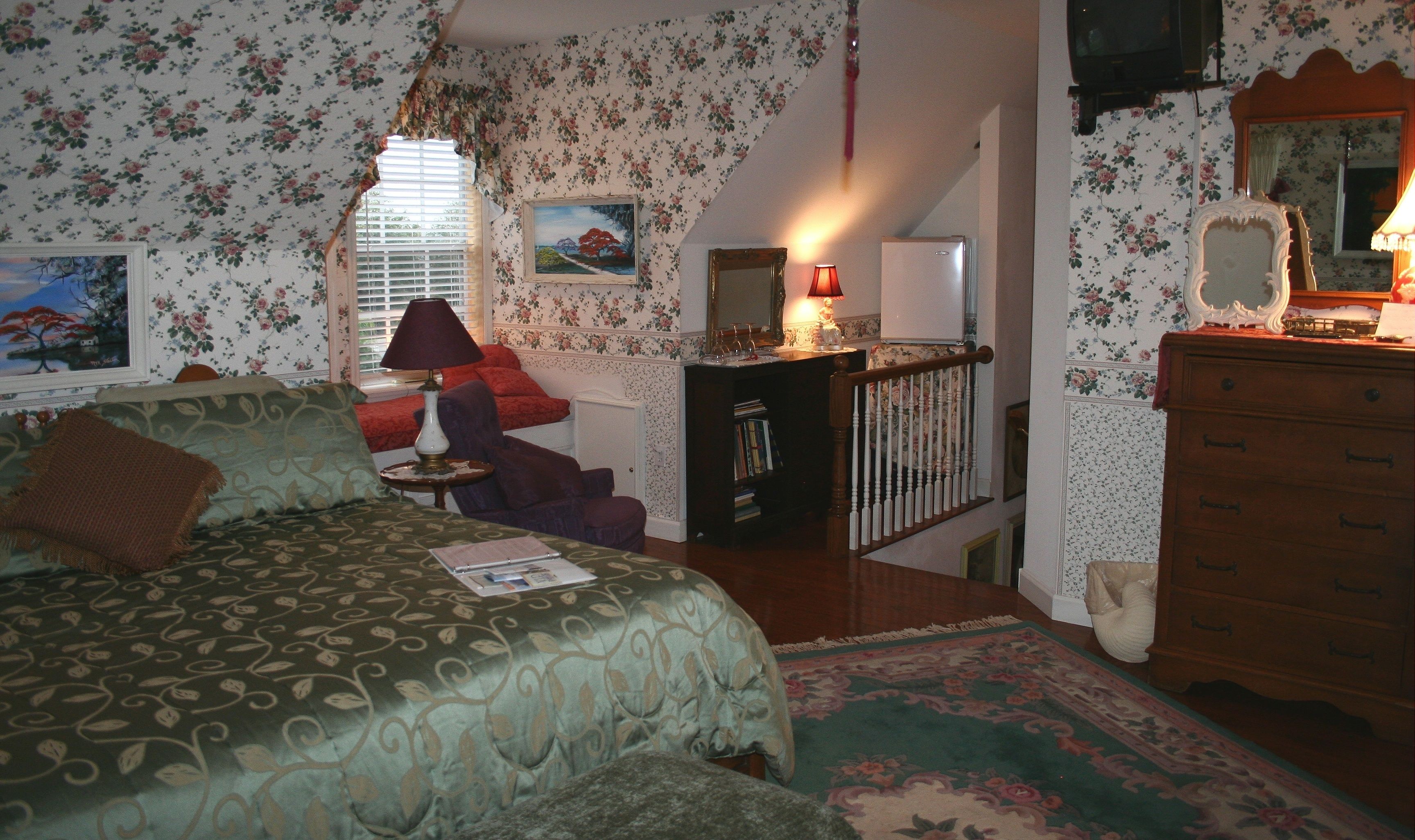 A bedroom with floral wallpaper, a bed with patterned bedding, a dresser, a desk with a lamp, and a window with blinds.
