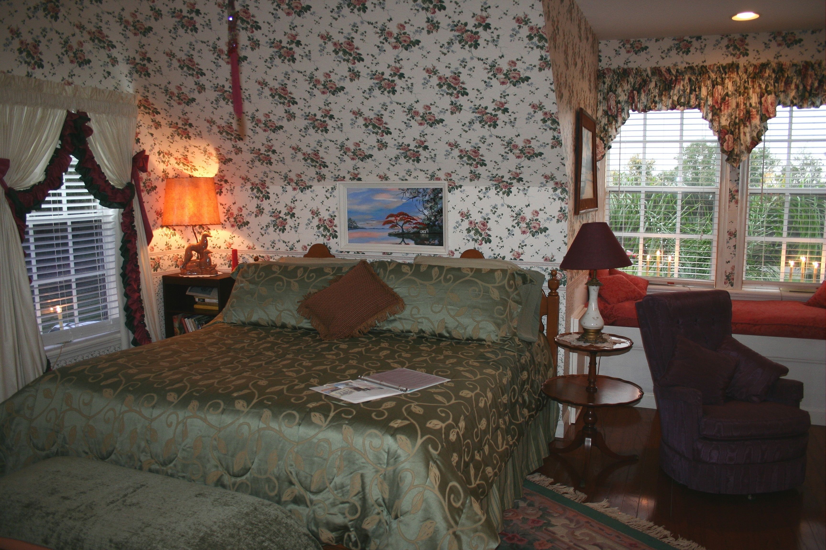 A bedroom with a bed, a nightstand, a chair, a window with curtains, a lamp, and a painting on the wall.