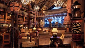 A spacious lobby with high ceilings, wooden beams, and a large window offering a view of the sky. Furnishings include wicker chairs, tables, and decorative lamps.