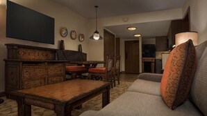 A living room with a sofa, a wooden coffee table, a dining table with chairs, a flat-screen TV mounted on the wall, and a pendant light.