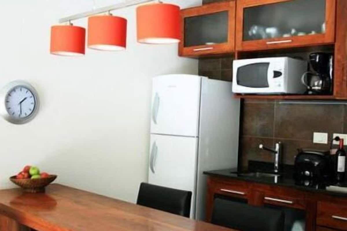 A kitchen with a white refrigerator, a microwave, and a coffee maker on the counter. There is a clock on the wall and a fruit bowl on the table.