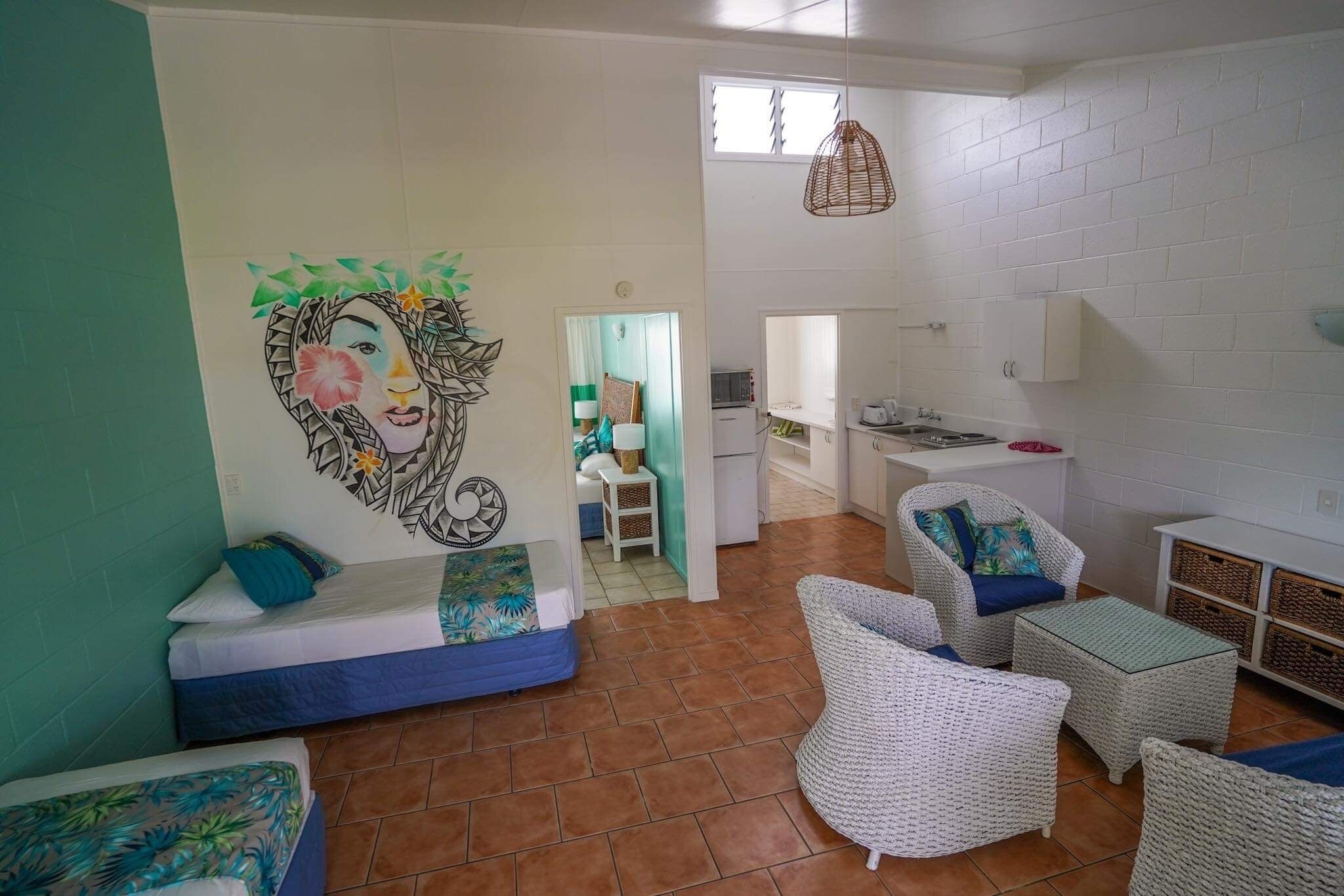 A room with a bed, a sofa, wicker furniture, a kitchen area, and a mural on the wall.