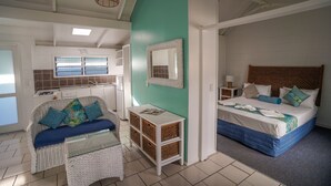 Ein Zimmer mit Bett, einer Couch, einem kleinen Tisch und einem Korbstuhl. Es gibt ein Fenster mit Jalousien, einen Spiegel an der Wand und eine Deckenleuchte.