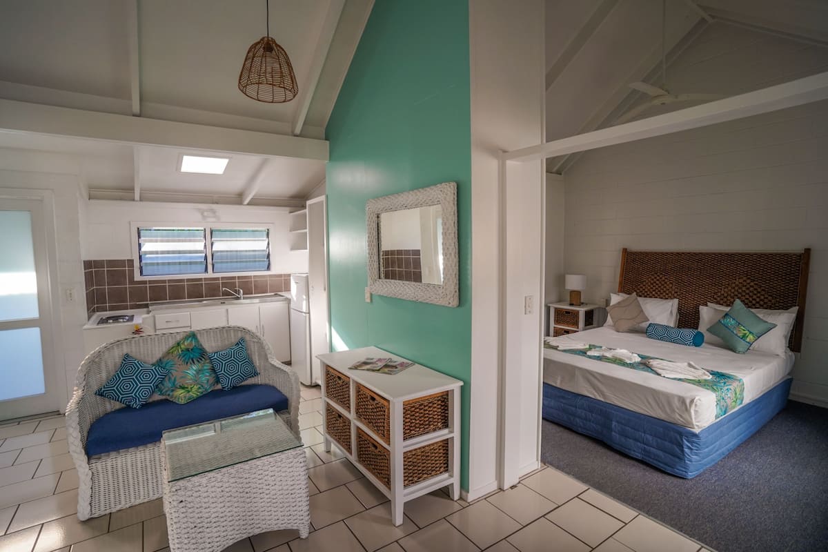 A room with a bed, a sofa, a small table, and a wicker chair. There is a window with blinds, a mirror on the wall, and a ceiling light fixture.