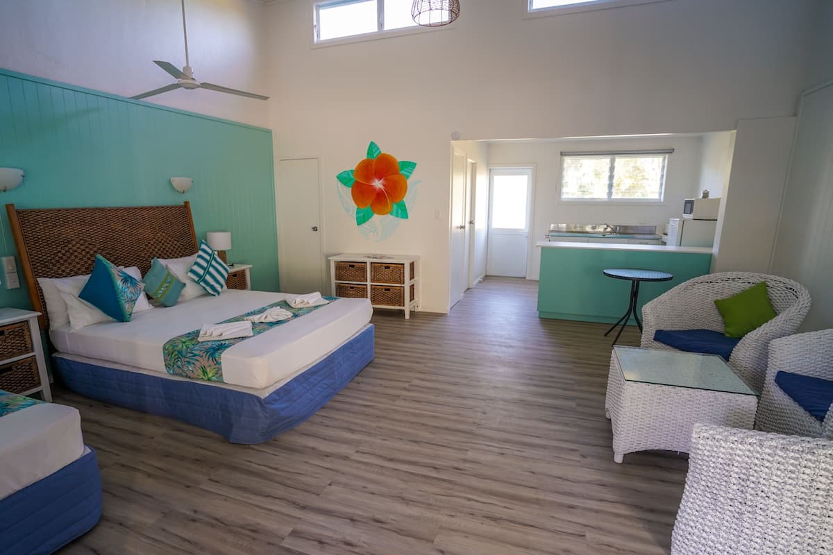 A spacious room with a large bed, a sitting area with wicker furniture, a kitchenette, and a ceiling fan.