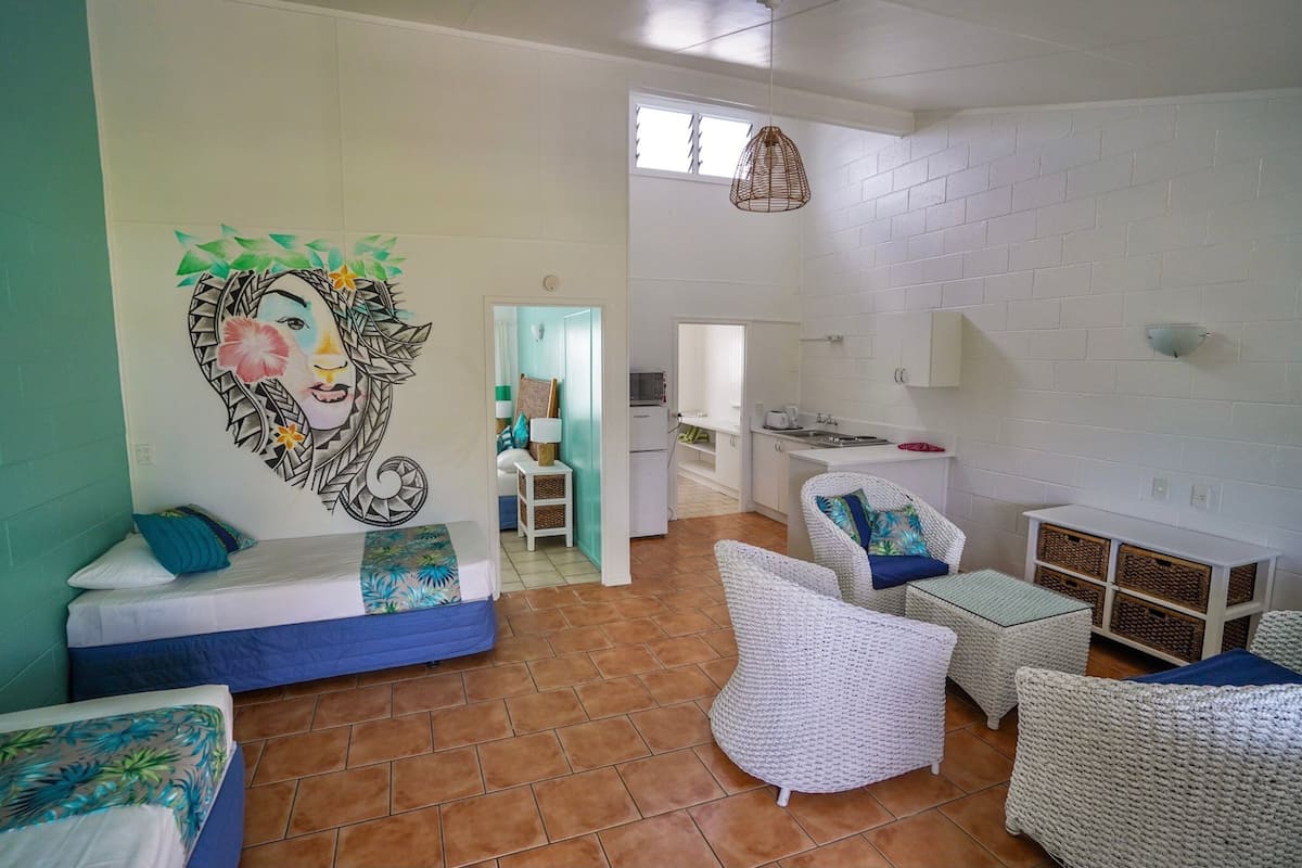 A room with a bed, white wicker furniture, a kitchen area, and a mural on the wall.