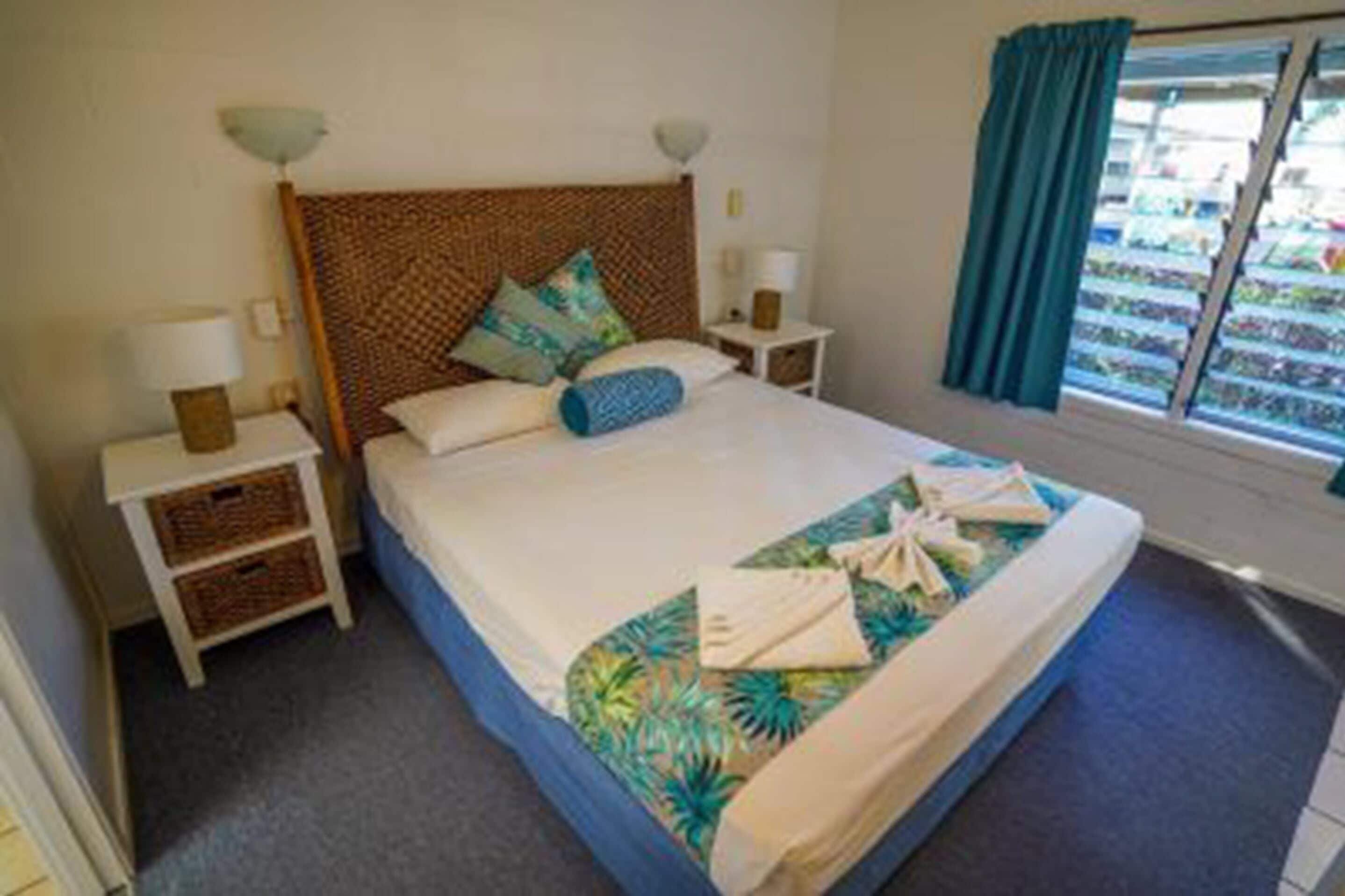 A neatly made bed with a tropical-themed bedspread, two bedside tables with lamps, and a view of a balcony with curtains.