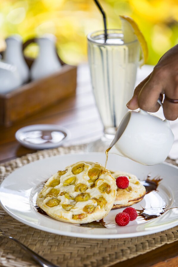 Una fuente de tortitas con sirope y frambuesas, una jarra de leche y un vaso de té helado sobre una mesa.
