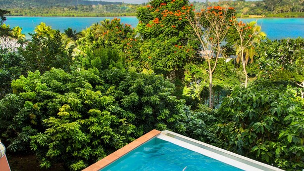 A swimming pool with a wooden deck, lounge chairs, and a person swimming. - Poppys On The Lagoon (Port Vila)