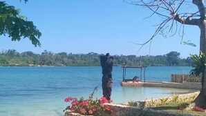 A person standing on a stone platform by a clear blue lake, overlooking a dock and a sandy beach. - Poppys On The Lagoon (Port Vila)