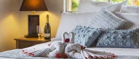 A neatly made bed with a towel sculpture of two swans, a bottle of wine, and a glass on a bedside table.