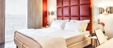 A modern hotel room with a large bed, a bedside table with a lamp, and a view of the outside through a large window.