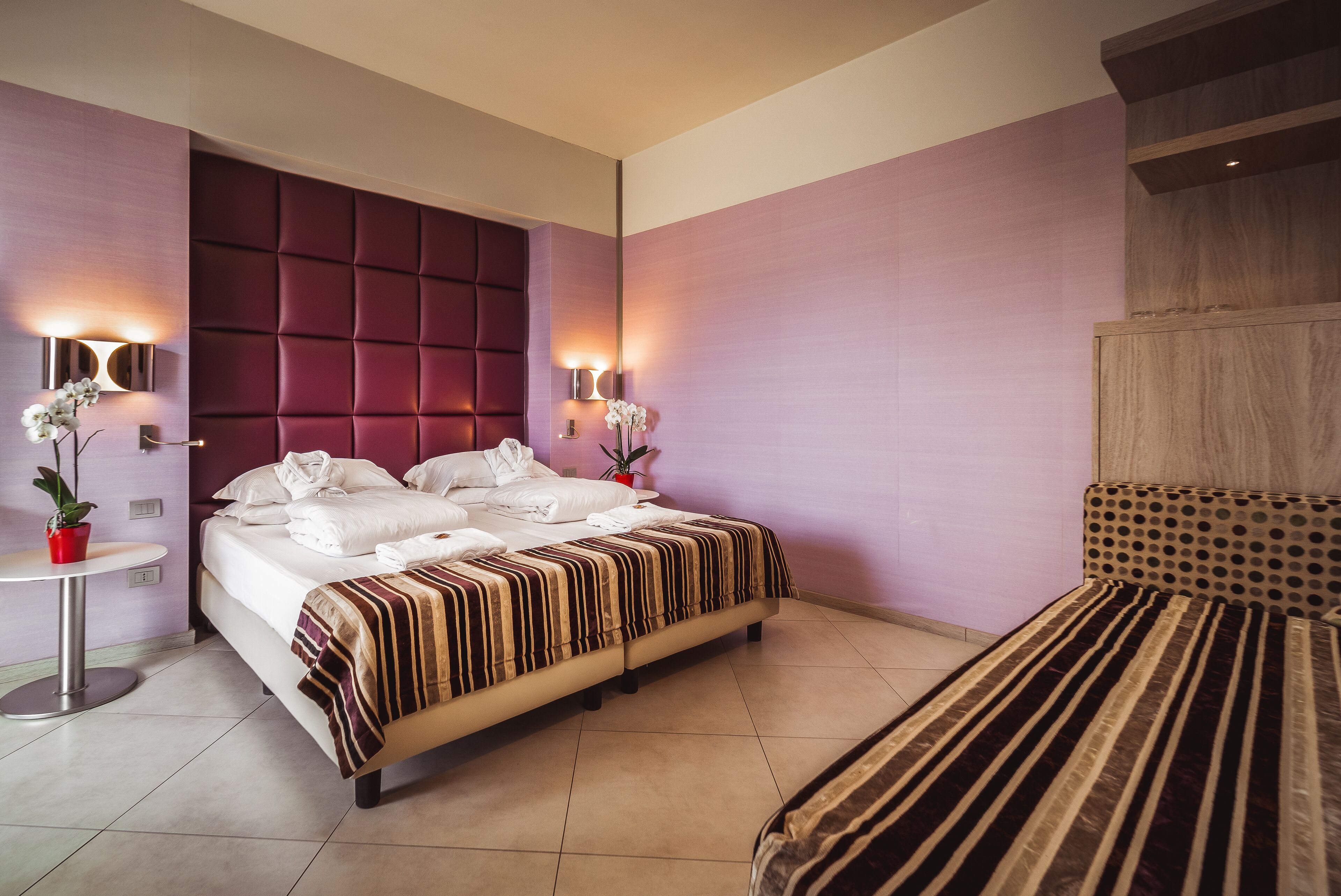 A modern hotel room with a large bed, a smaller bed, a round table with a potted plant, and a wall-mounted light fixture.