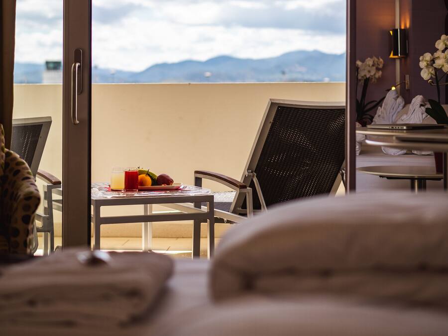 Ein Hotelzimmer mit Balkon, einem Esstisch mit Obst und einem Glas Saft sowie einem Bett mit Kissen.