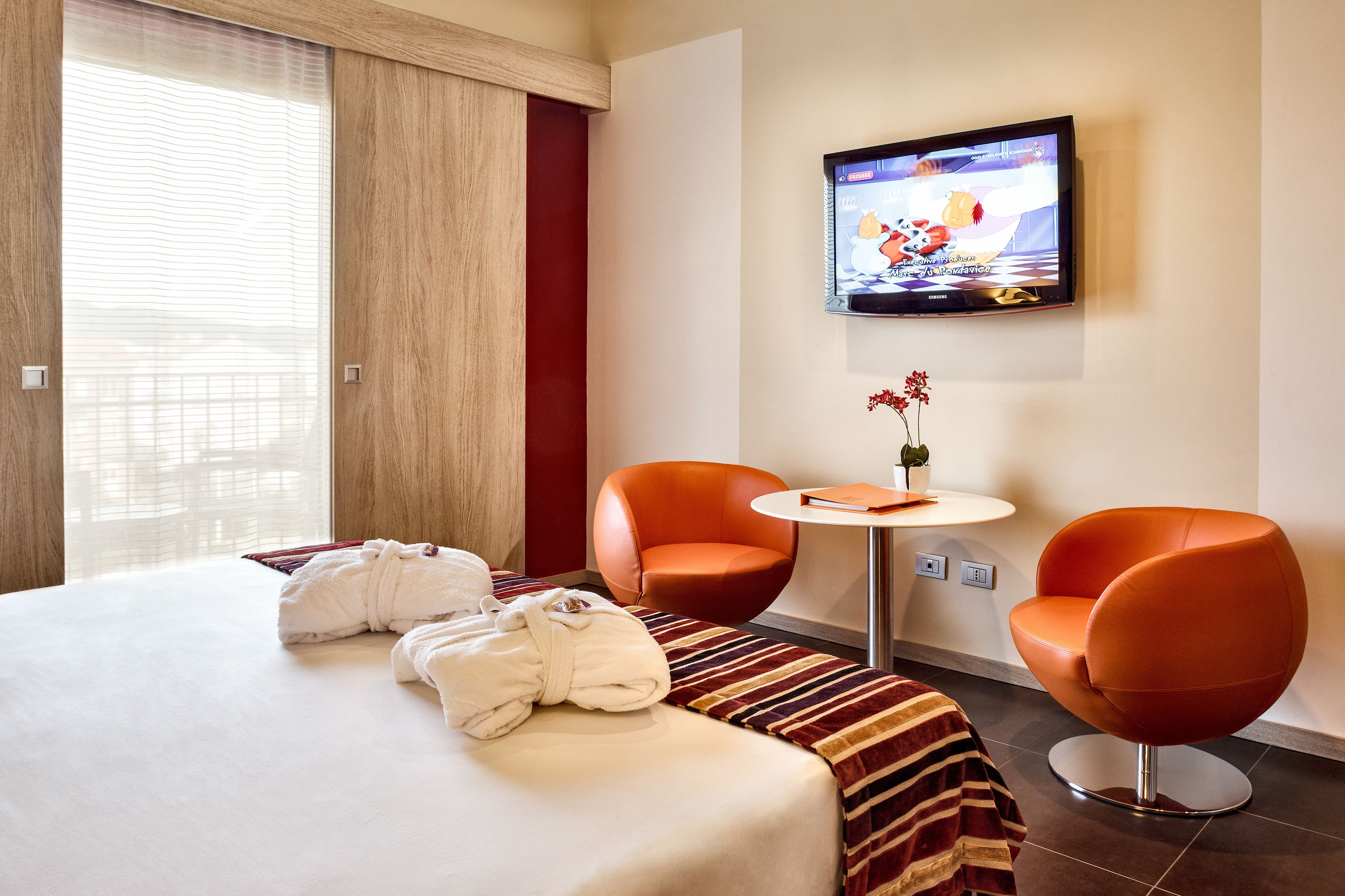 A hotel room with a bed, two orange chairs, a small table with a vase, and a TV on the wall.