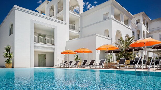 Un área junto a la piscina con sillones de descanso y sombrillas, una piscina azul cristalina y un edificio blanco con balcones.