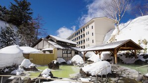 A traditional Japanese onsen with a wooden bathhouse, a large building with multiple windows, and a clear blue sky. - Manza Kogen Hotel (Tsumagoi)