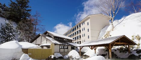 A traditional Japanese onsen with a wooden bathhouse, a large building with multiple windows, and a clear blue sky.