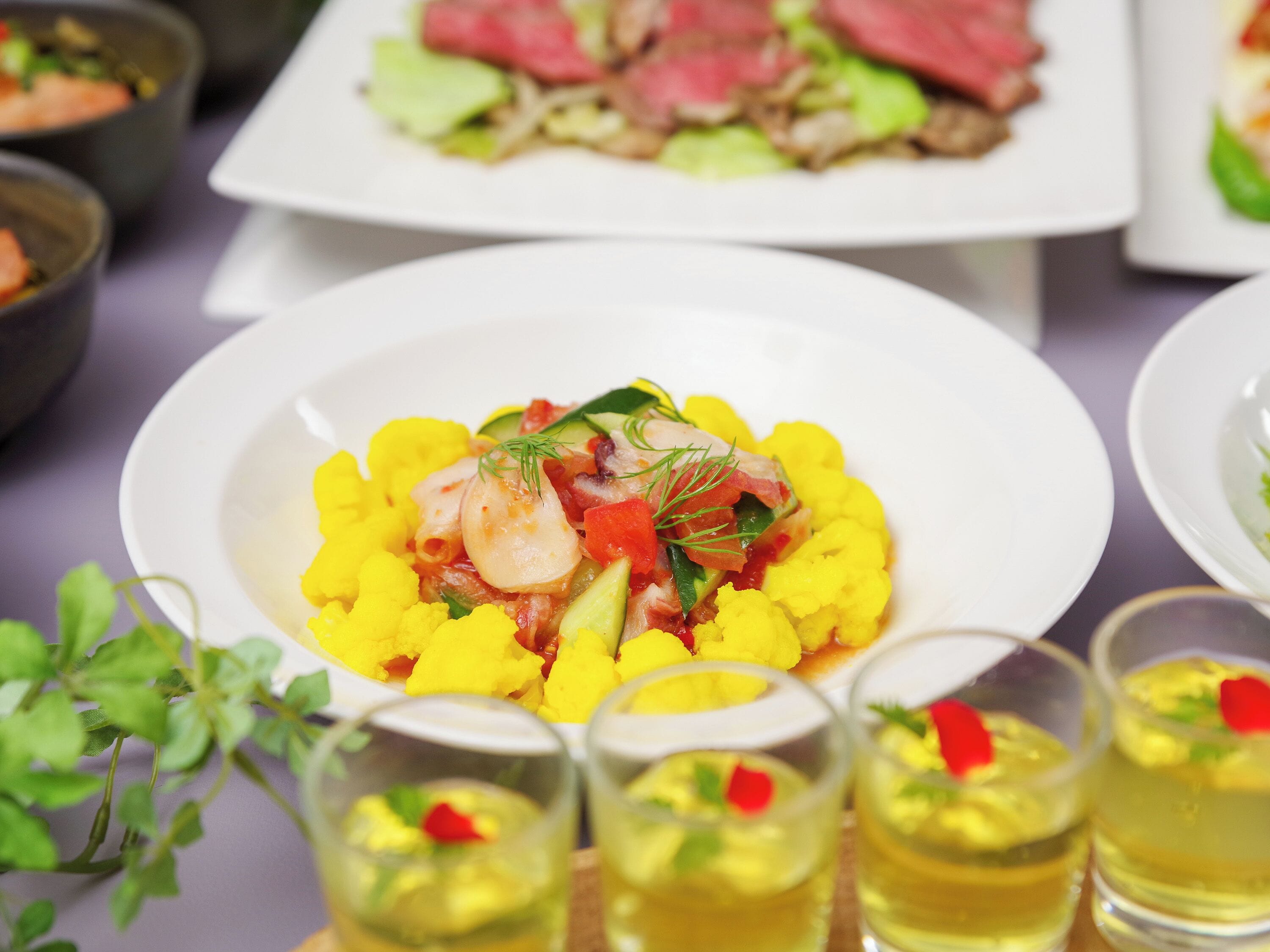 A dining table with various dishes, including a plate of yellow cauliflower and a plate of steak with vegetables. There are also small glasses with yellow drinks and a plant in the foreground.