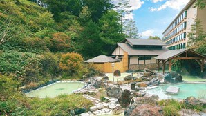 A spa area with multiple hot tubs surrounded by rocks and greenery, a wooden pavilion, and a building with a sloped roof. - Manza Kogen Hotel (Tsumagoi)