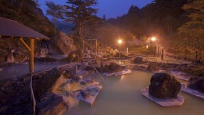 A hot spring area with a wooden deck, rocky terrain, and a metal railing. - Manza Kogen Hotel (Tsumagoi)