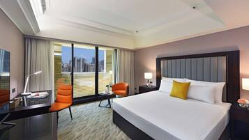 A hotel room with a large bed, two chairs, a desk, and a view of a cityscape through large windows.