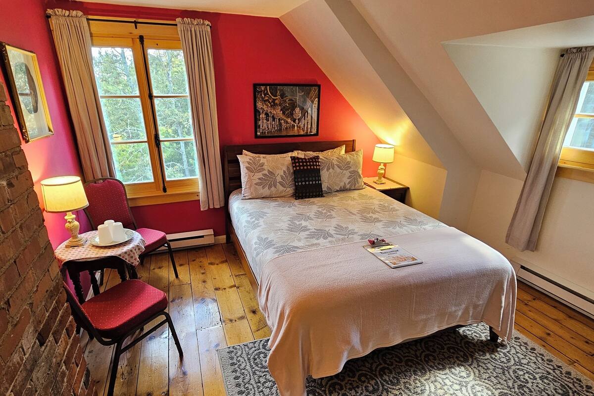 A cozy bedroom with a bed, a chair, a small table with cups, and a painting on the wall.