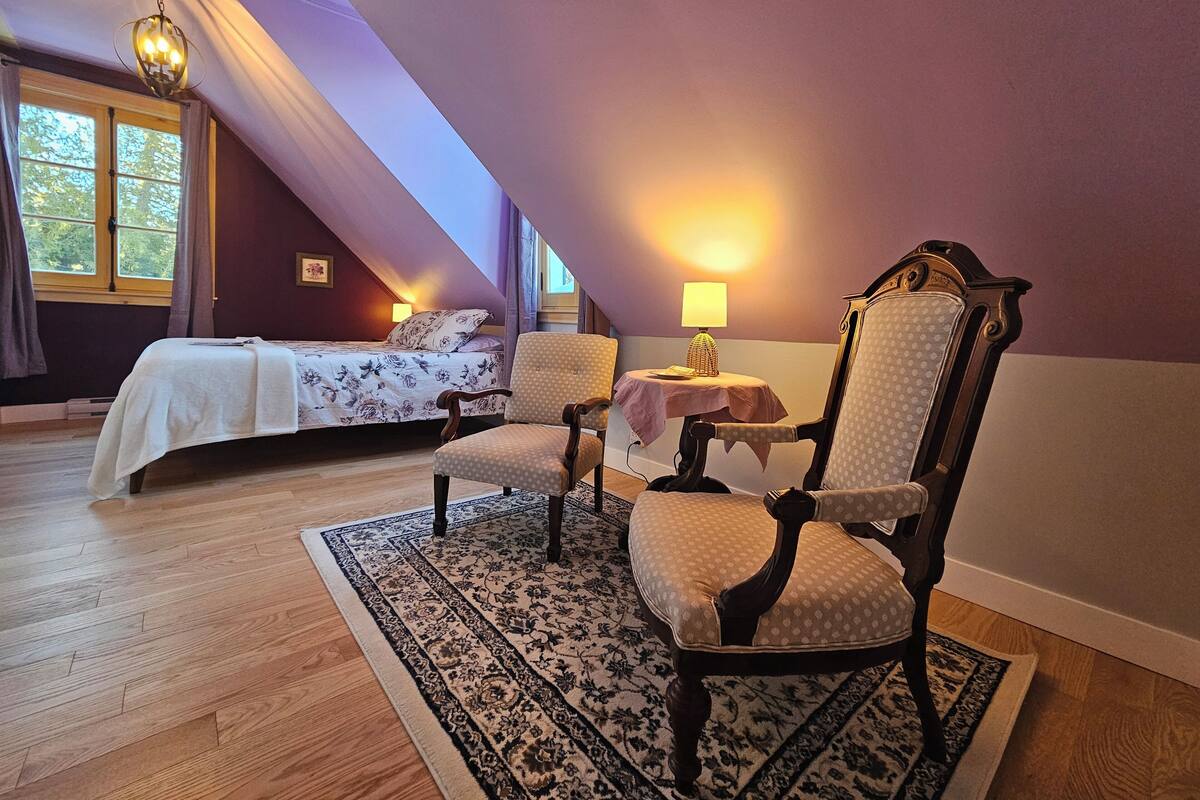 A bedroom with a bed, two chairs, a table, and a lamp.