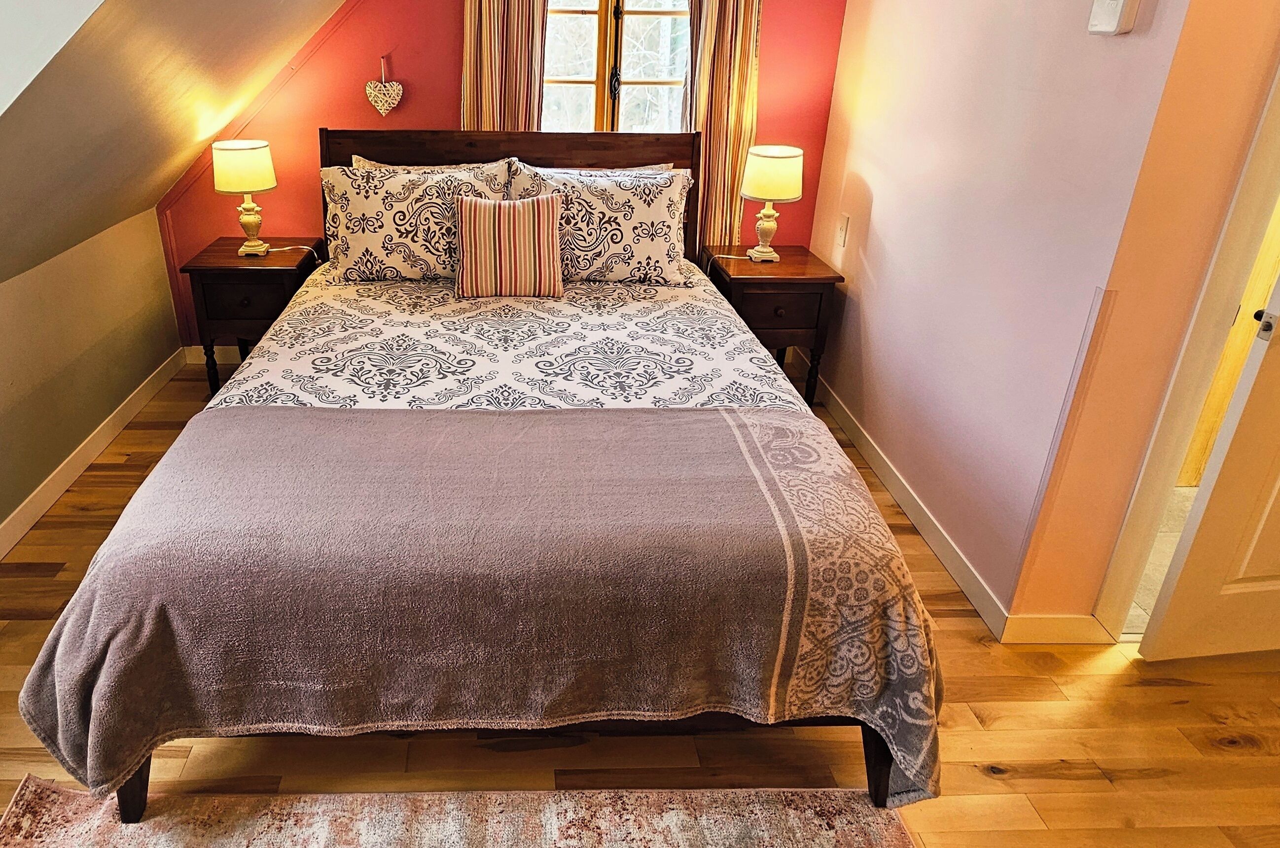 Un dormitorio con una cama grande, dos mesitas de noche, una mesita auxiliar, una ventana con cortinas y una decoración con forma de corazón en la pared.