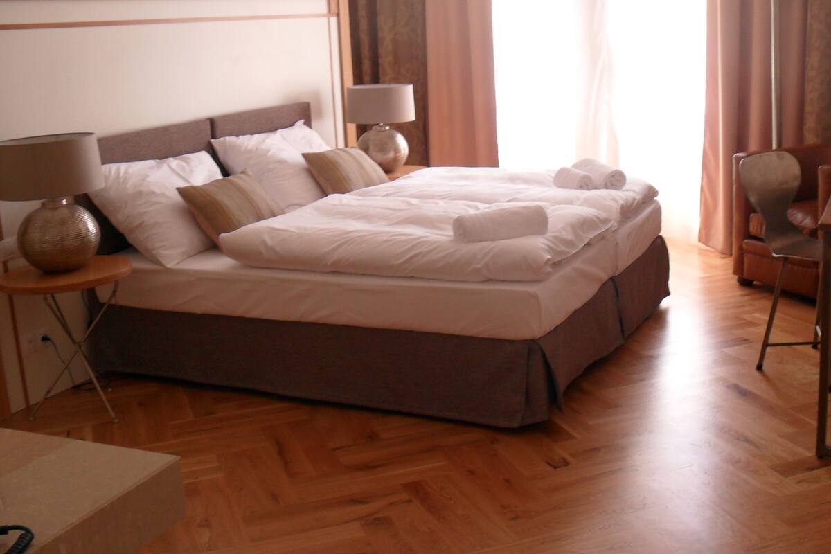 A hotel room with a large bed, two bedside tables with lamps, a wooden floor, a desk with a chair, and a window with curtains.
