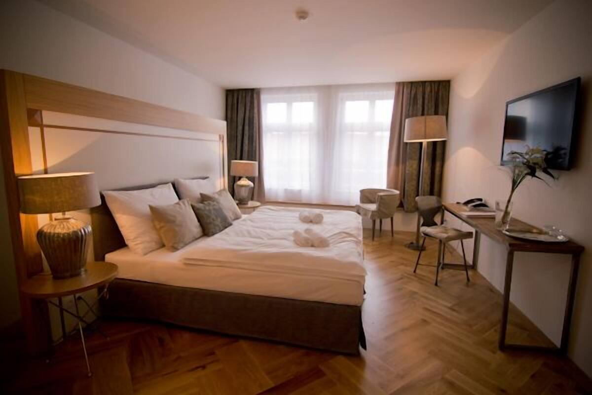 A hotel room with a large bed, two bedside tables with lamps, a desk with a chair, a flat-screen TV, and a large window with curtains.
