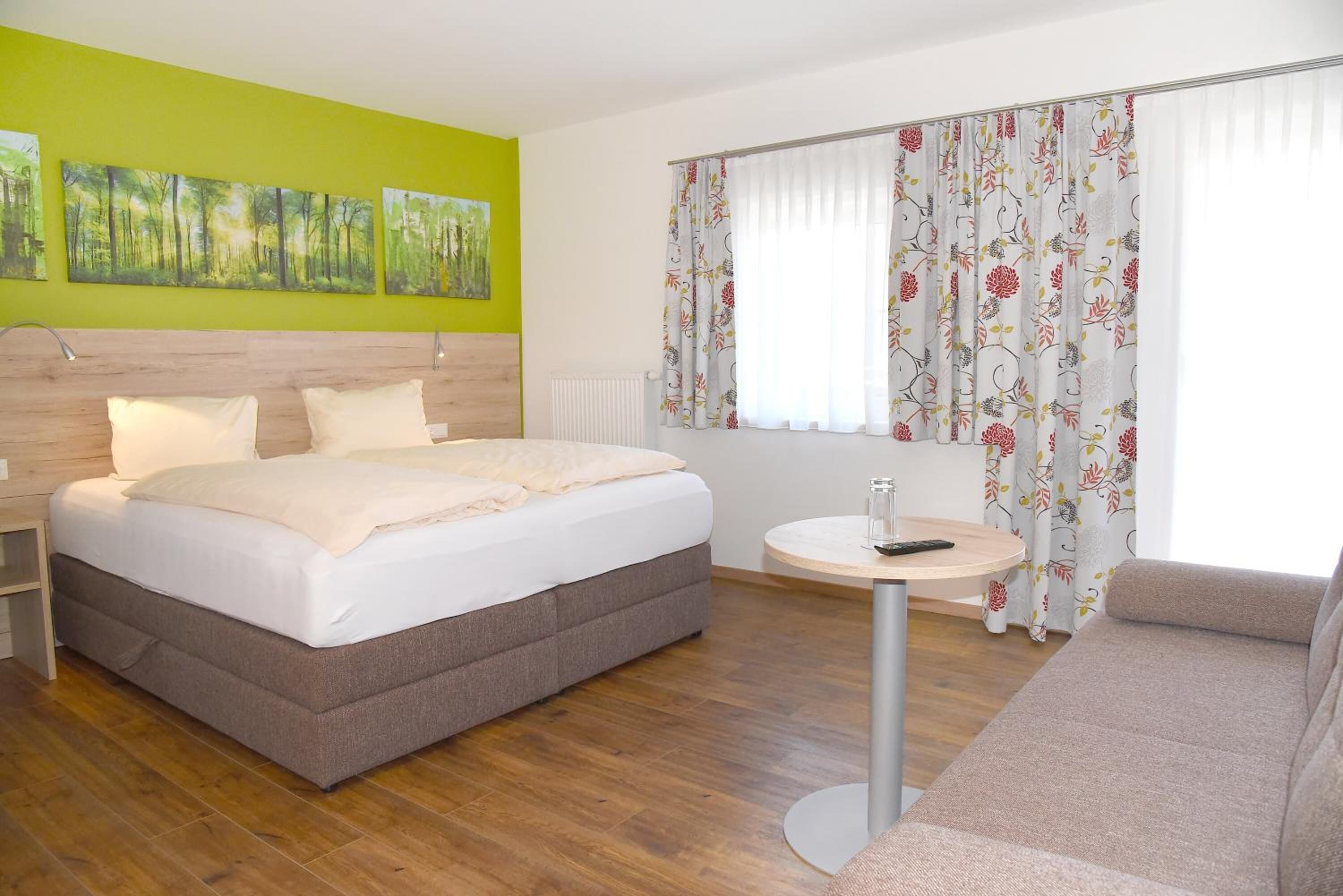 A hotel room with a bed, a round table, a sofa, and a window with floral curtains.