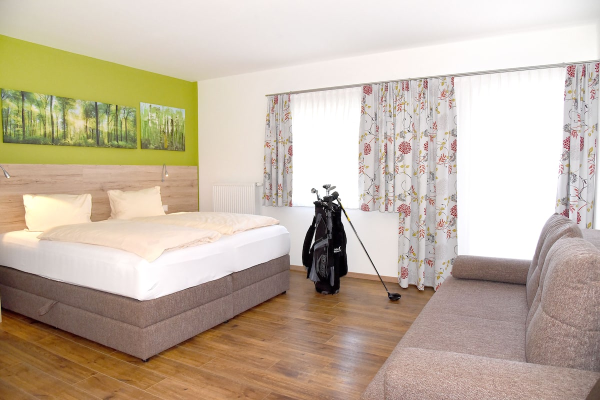 A hotel room with a bed, a sofa, a golf bag, and a window with floral curtains.