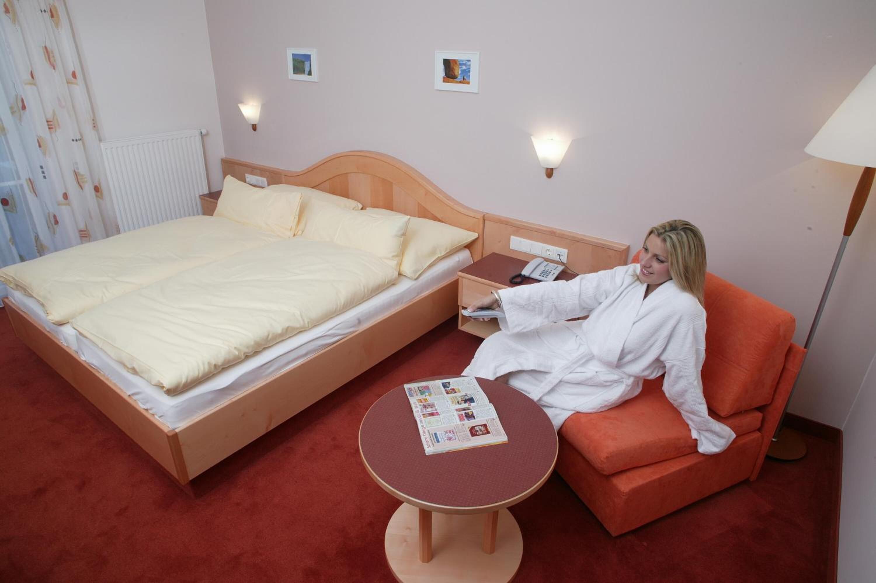 Een hotelkamer met een groot bed, een nachtkastje met een lamp, een klein rond tafeltje met een tijdschrift, en een rode fauteuil.