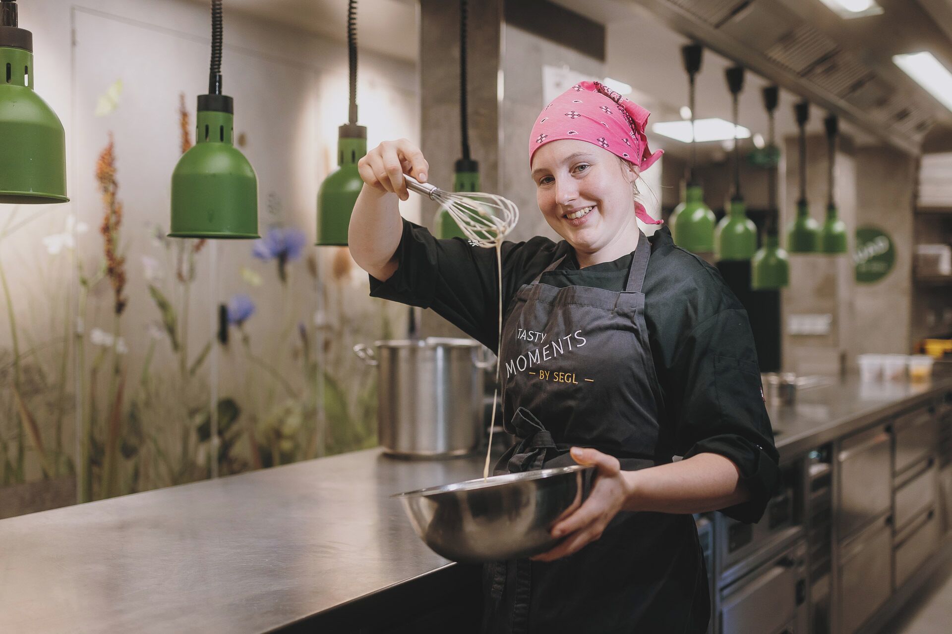 Une personne se trouve dans une cuisine, tenant un fouet et un saladier. Des guirlandes lumineuses vertes sont suspendues au plafond et le plan de travail est en acier inoxydable.