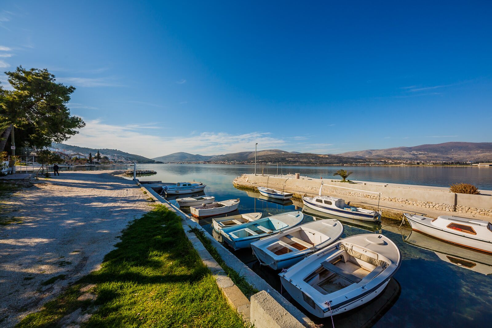 A marina with several small boats docked along the shore, a clear sky, and a distant view of hills.