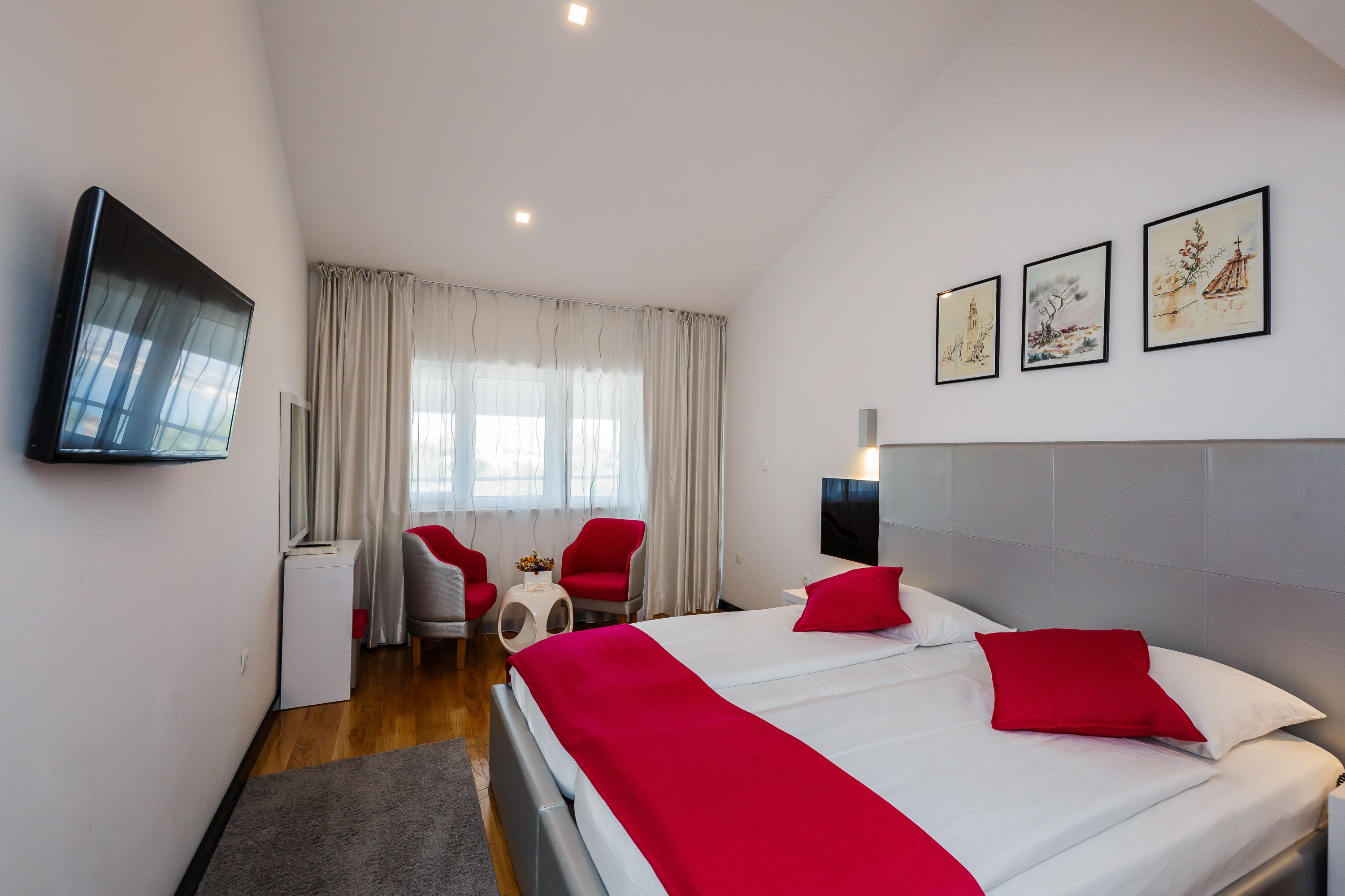 A hotel room with a large bed, red pillows, a flat-screen TV, a desk with a chair, and artwork on the wall.