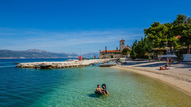 Una playa con aguas transparentes, un flotador con forma de cisne y una iglesia al fondo.