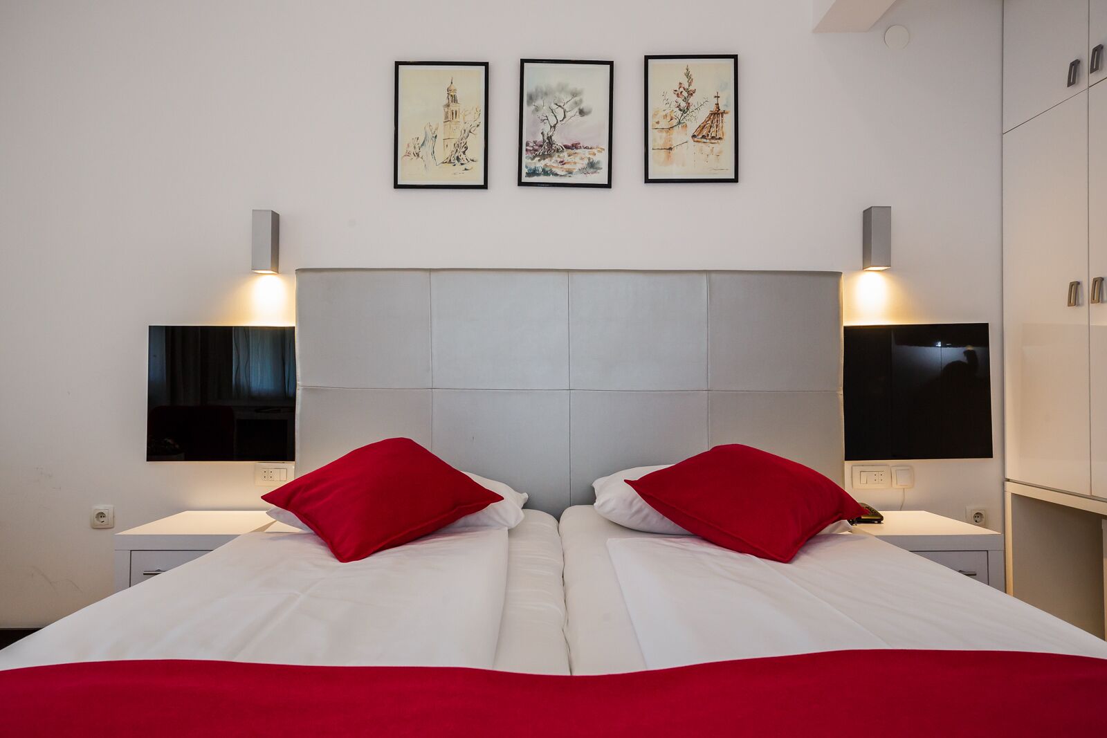 A hotel room with a large bed, two red pillows, a white headboard, and three framed artworks on the wall.