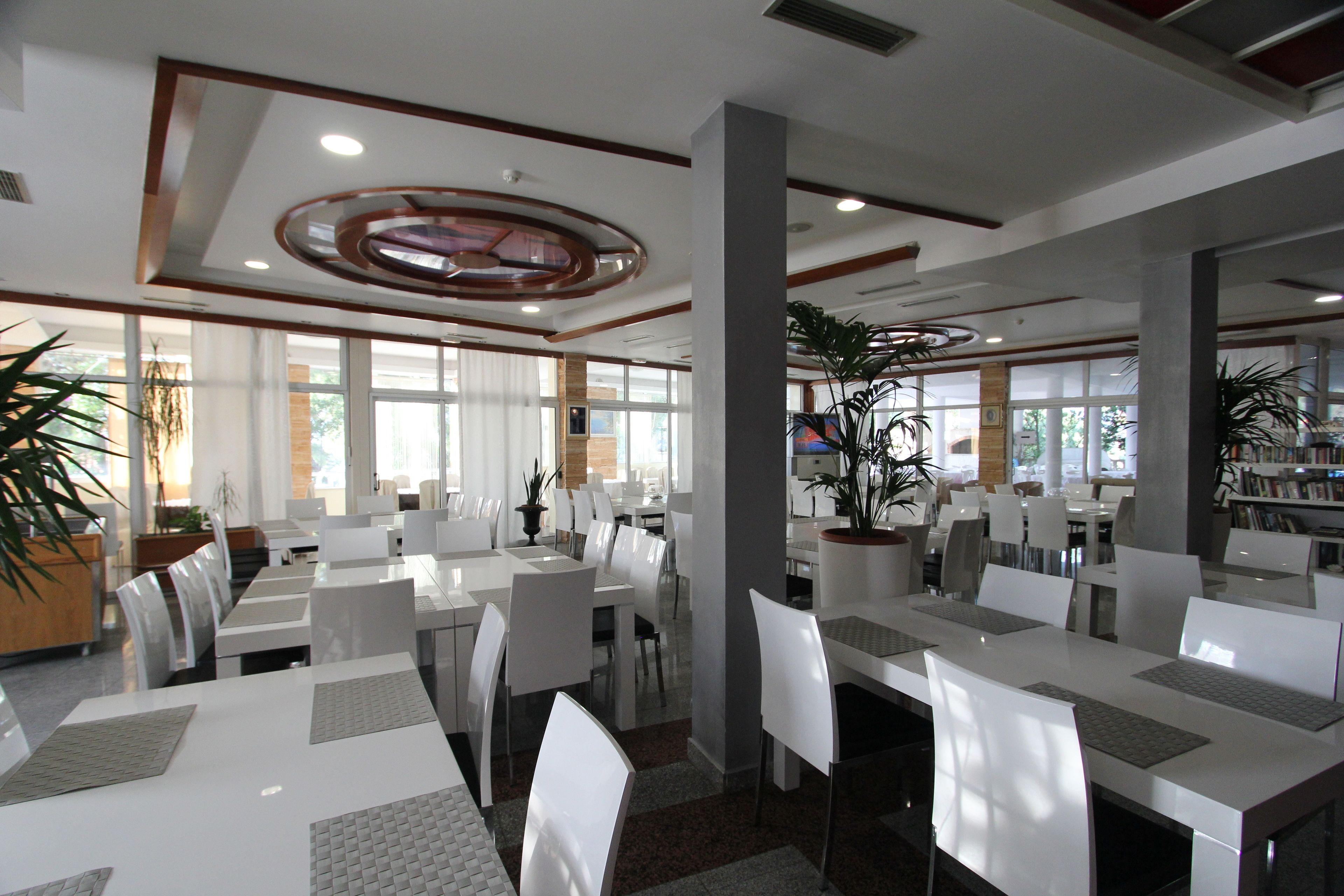 Een ruim restaurant met witte tafels en stoelen, grote ramen en potplanten.