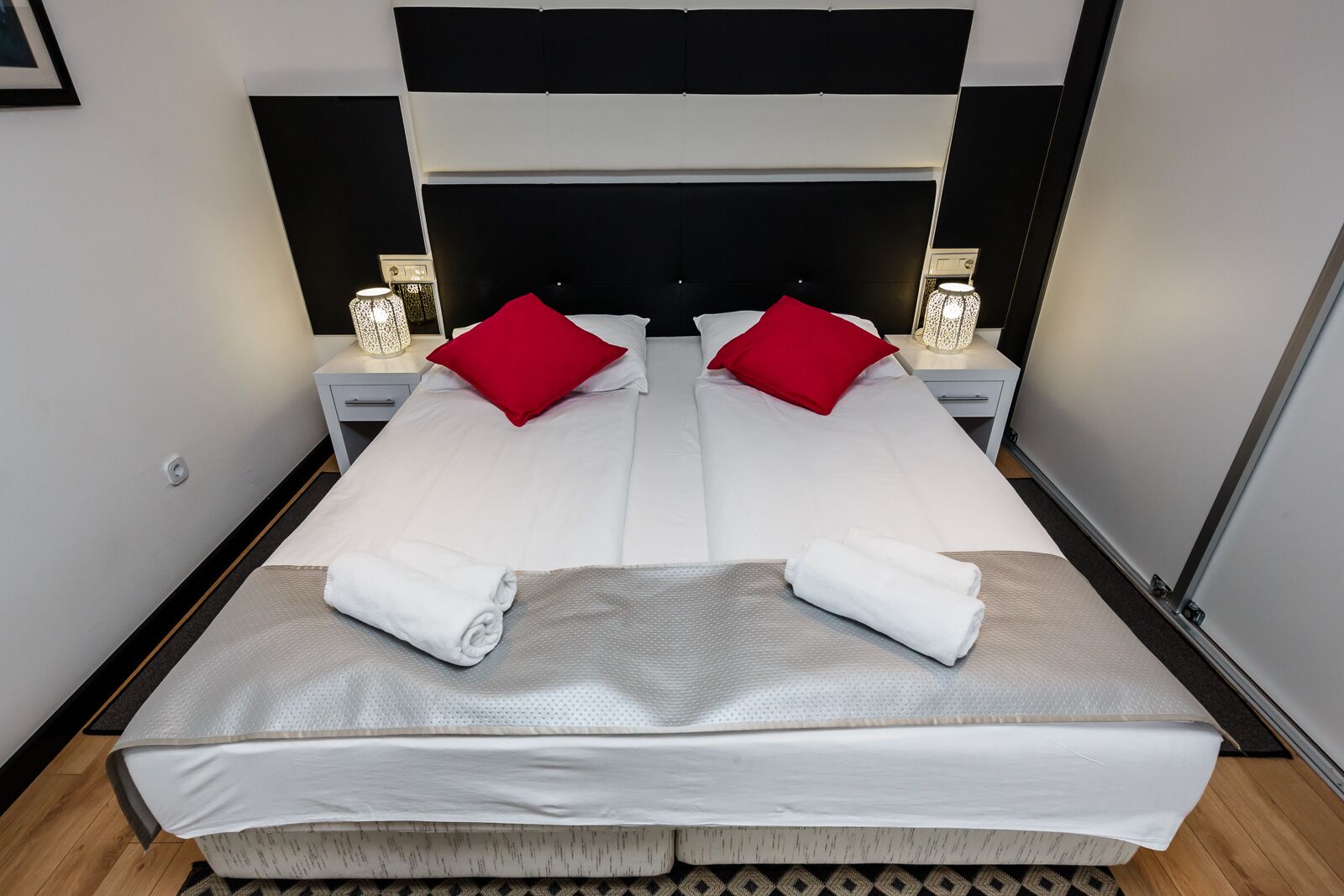 A double bed with white bedding and red pillows, two bedside tables with lamps, and a black headboard.