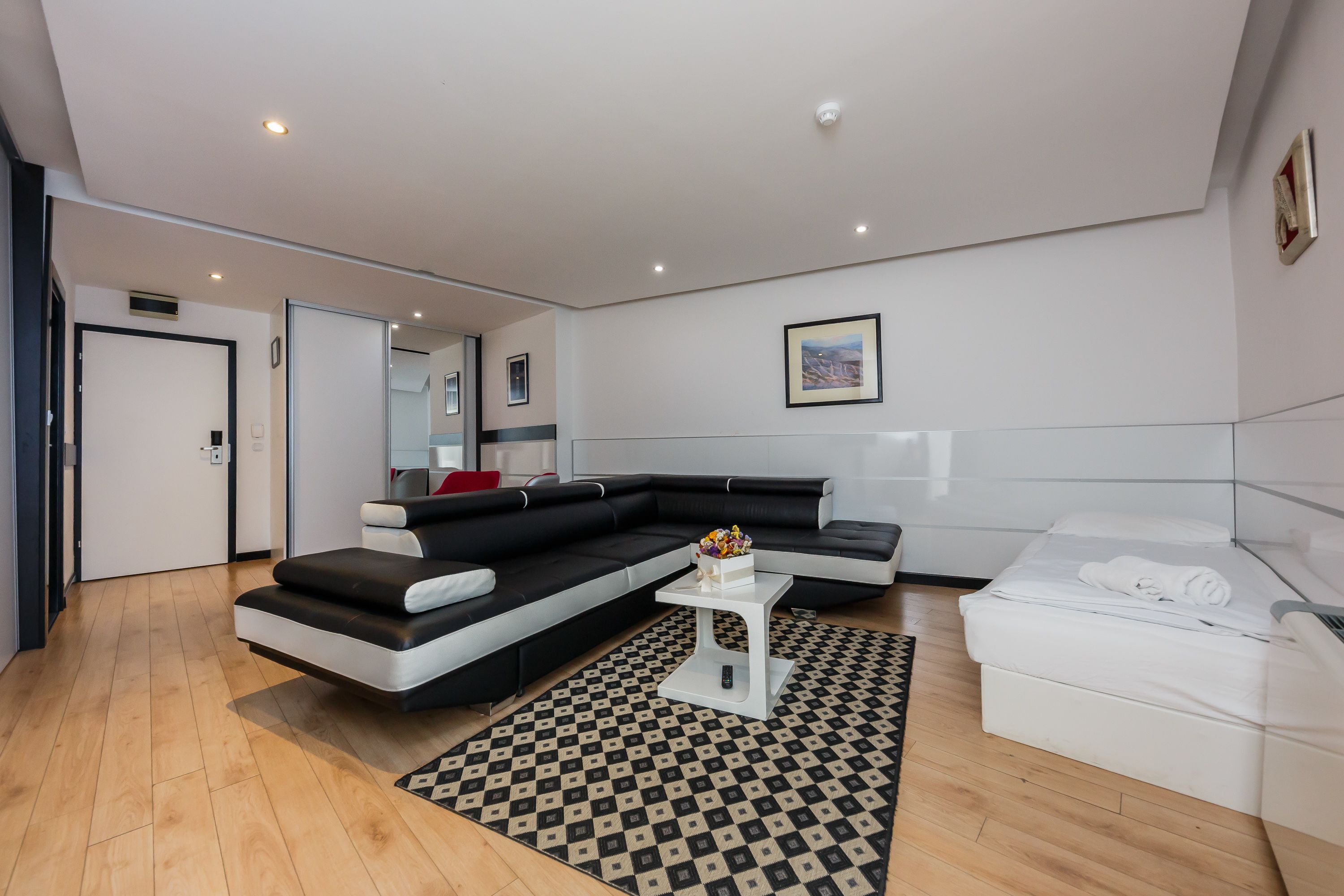 Ein modernes Wohnzimmer mit einem Ecksofa, einem weißen Couchtisch, einem Bett und einem gemusterten Teppich.