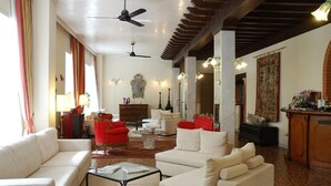 A spacious living room with a high ceiling, a ceiling fan, and a large window with curtains. There are several pieces of furniture including a sofa, armchairs, and a coffee table. - Hotel Valdor (Cavallino-Treporti)