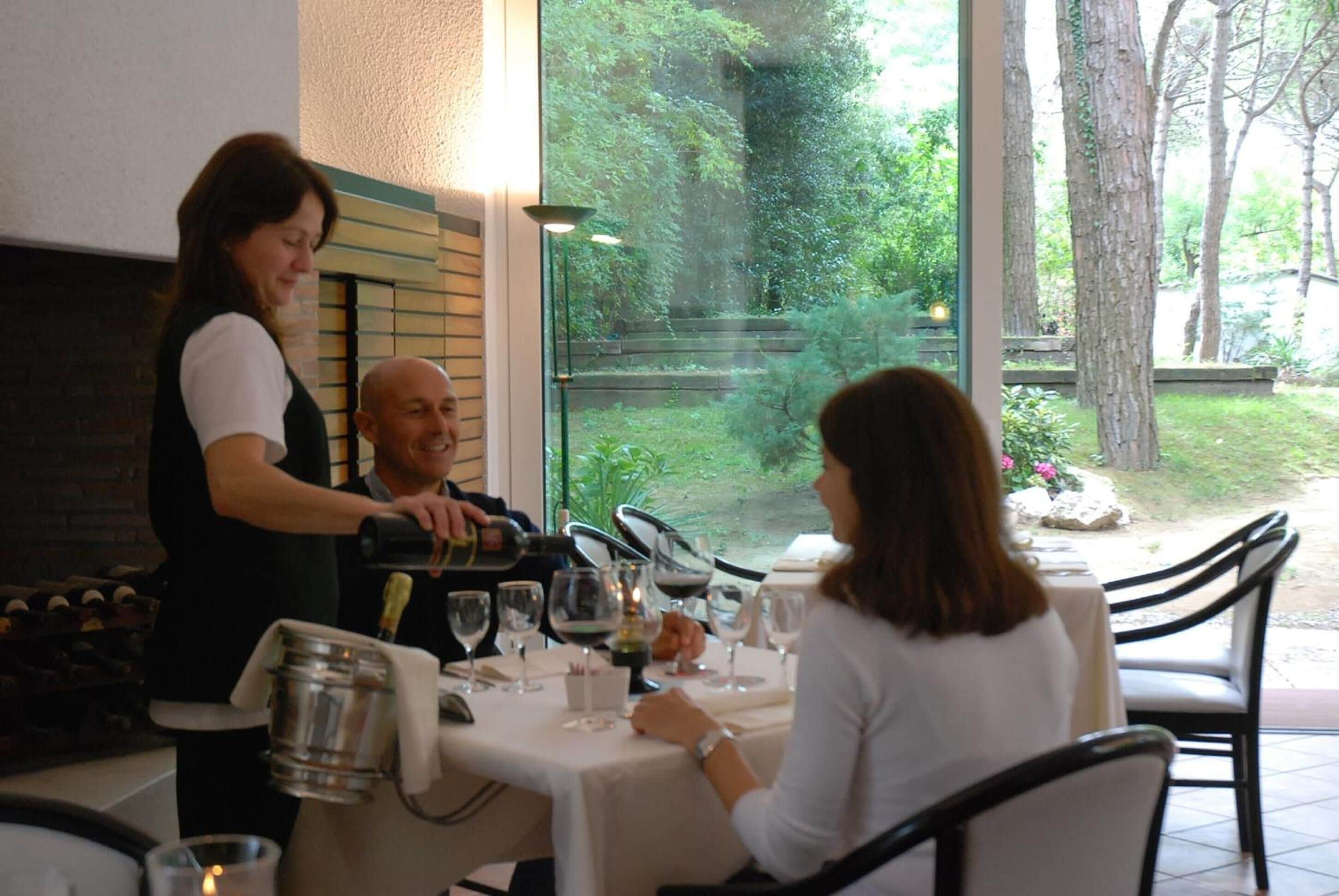A restaurant with a table set for two, a waiter serving, and a view of greenery outside.