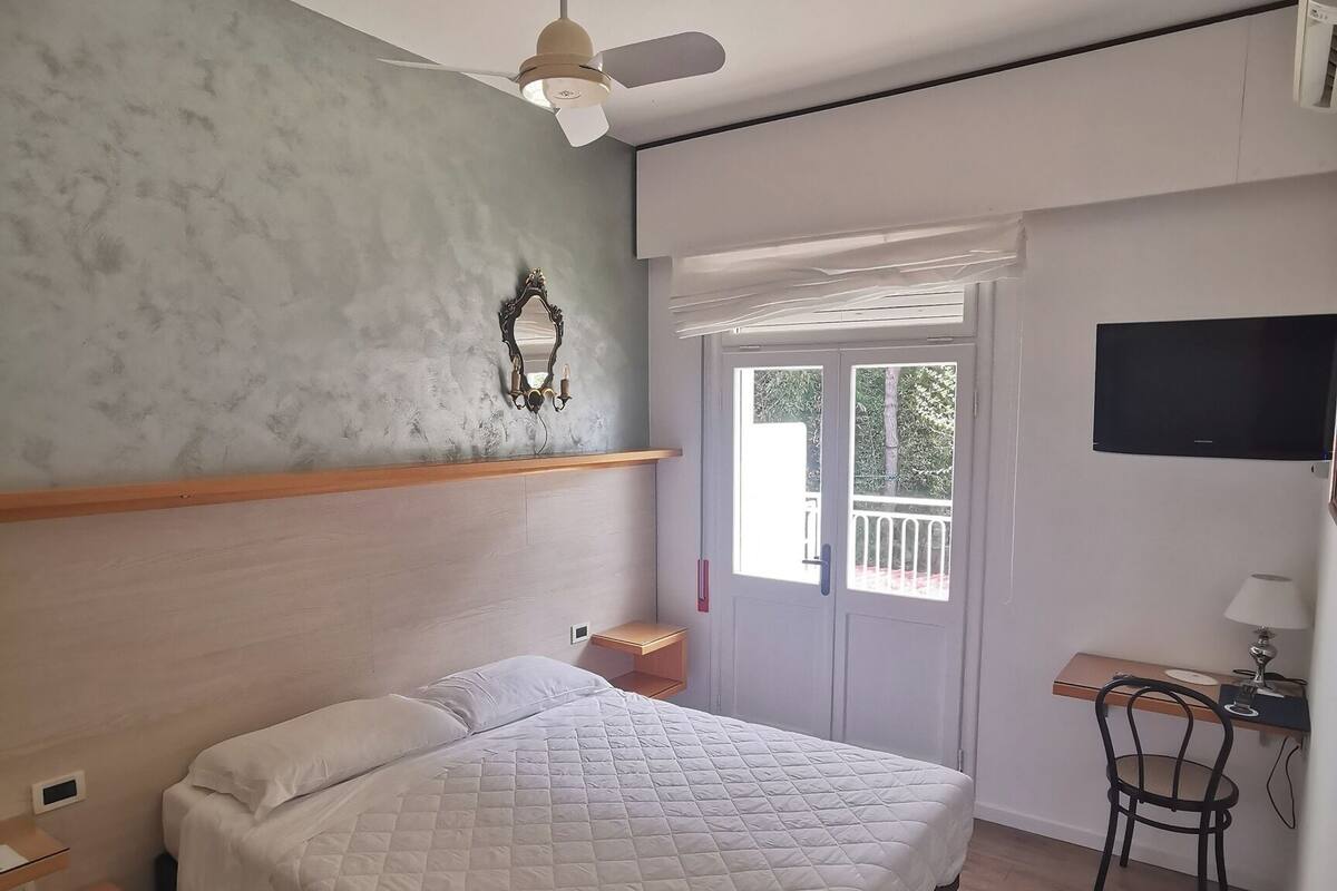 A bedroom with a bed, a ceiling fan, a small table with a chair, and a TV mounted on the wall.