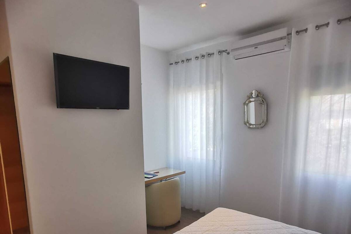 A bedroom with a bed, a TV mounted on the wall, a chair, and a window with curtains.