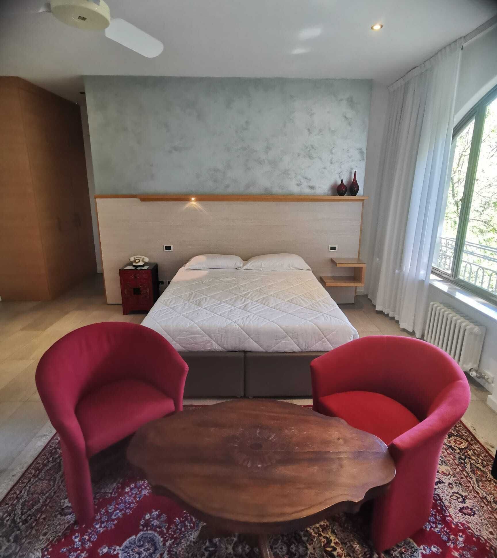 A bedroom with a bed, two red chairs, a wooden table, a nightstand, and a window with curtains.